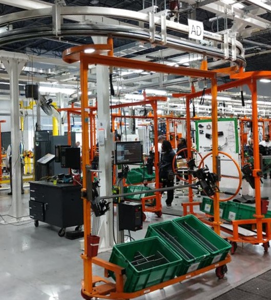 Industrial assembly line with orange frames, green bins, monitors, and workers in a factory.