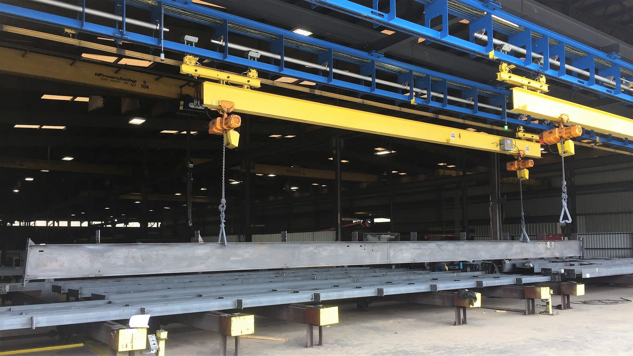 Yellow overhead cranes lifting a large metal beam inside an industrial workshop.