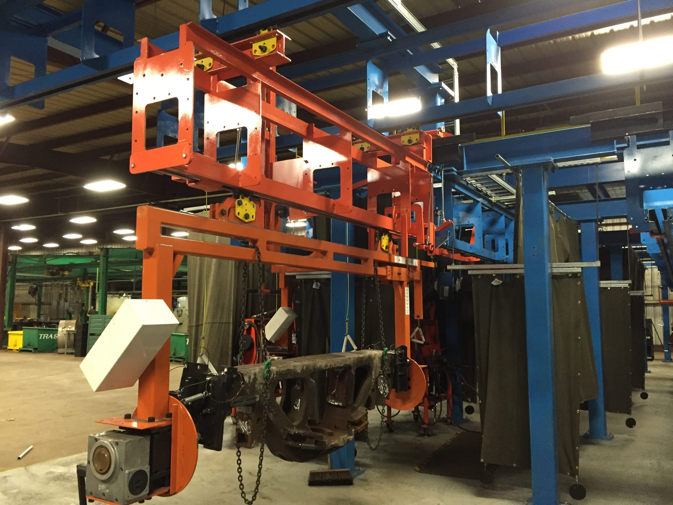 Industrial factory setup with orange metal frame and suspended dark metal component on blue conveyor system.