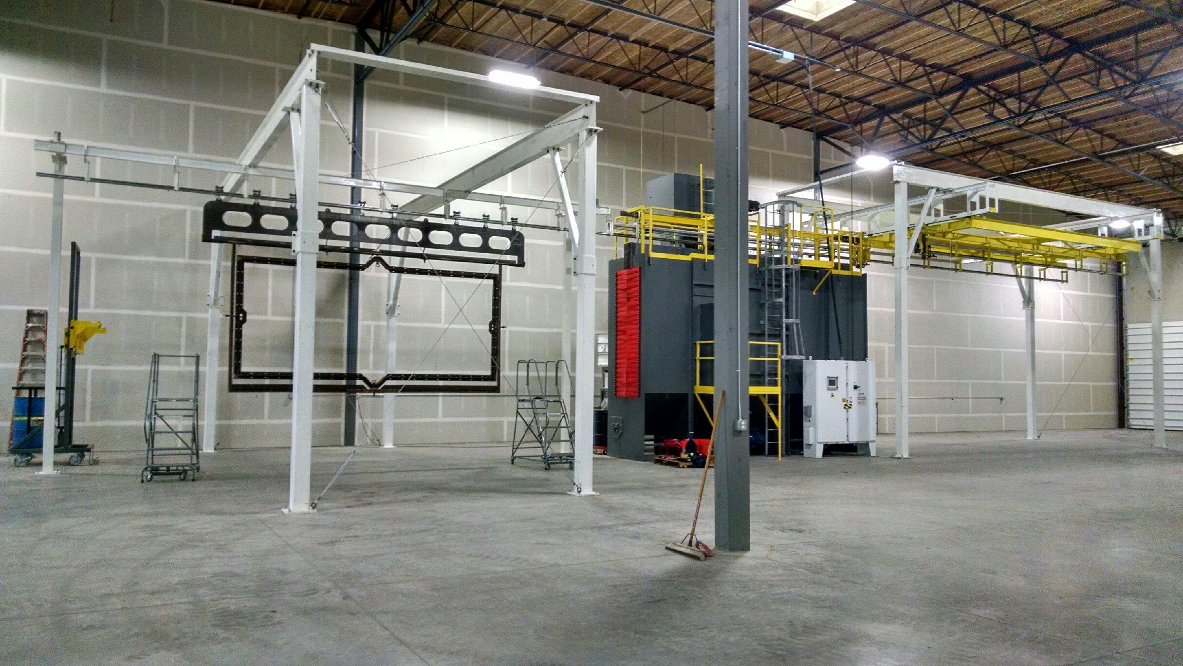 Industrial warehouse interior with machinery, metal frames, ladders, and broom on concrete floor.